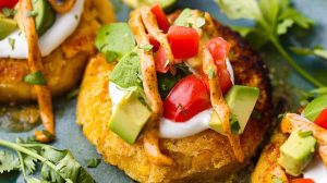 Angle shot of sweet corn tamale cakes plated and topped with fresh tomatoes, avocado, cilantro and sauce.