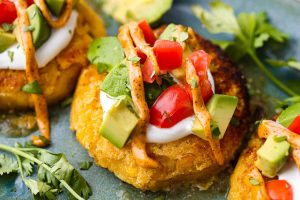 Angle shot of sweet corn tamale cakes plated and topped with fresh tomatoes, avocado, cilantro and sauce.