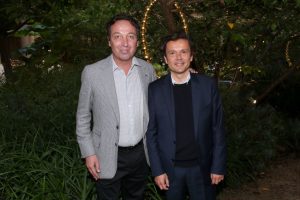 Two men in jackets stand in front of some foliage.